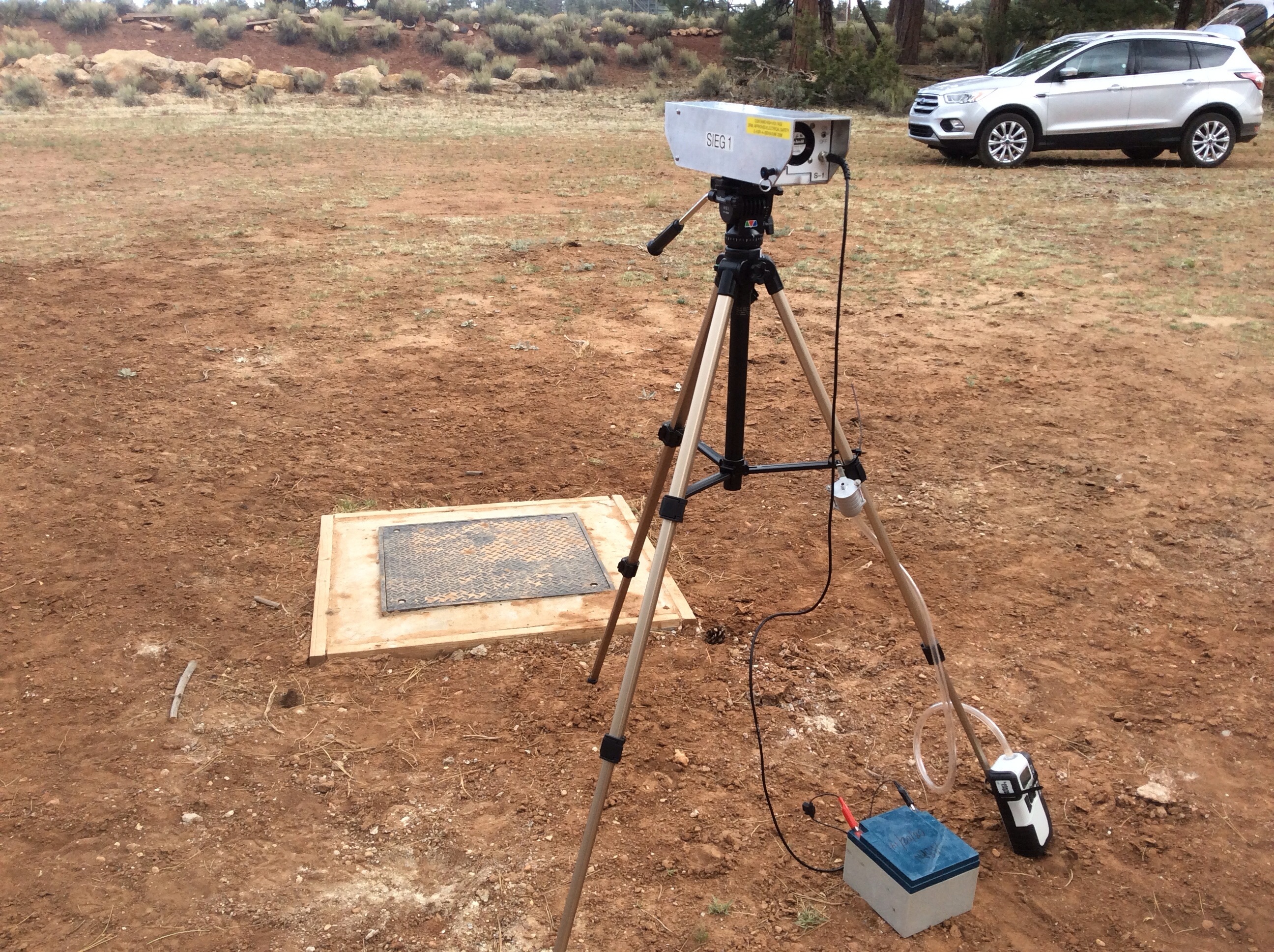  Tripod with active dust sampler on it pointed toward the mine site and attached to batteries.  Concrete and metal structure on the ground is groundwater well covering. 