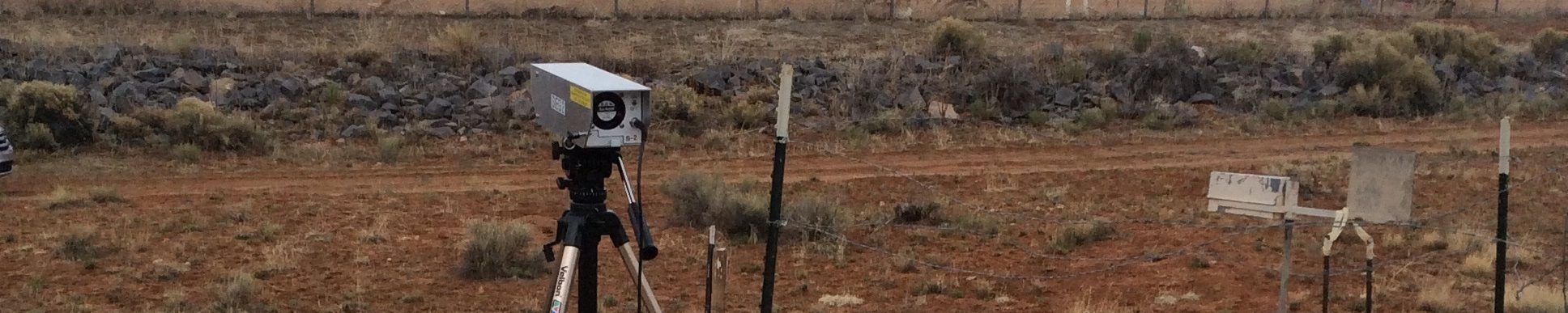  Active dust sampler sitting on top of tripod and attached to batteries. Head frame and fence of the Pinyon Plain Mine (formerly Canyon Mine) in the background. 
