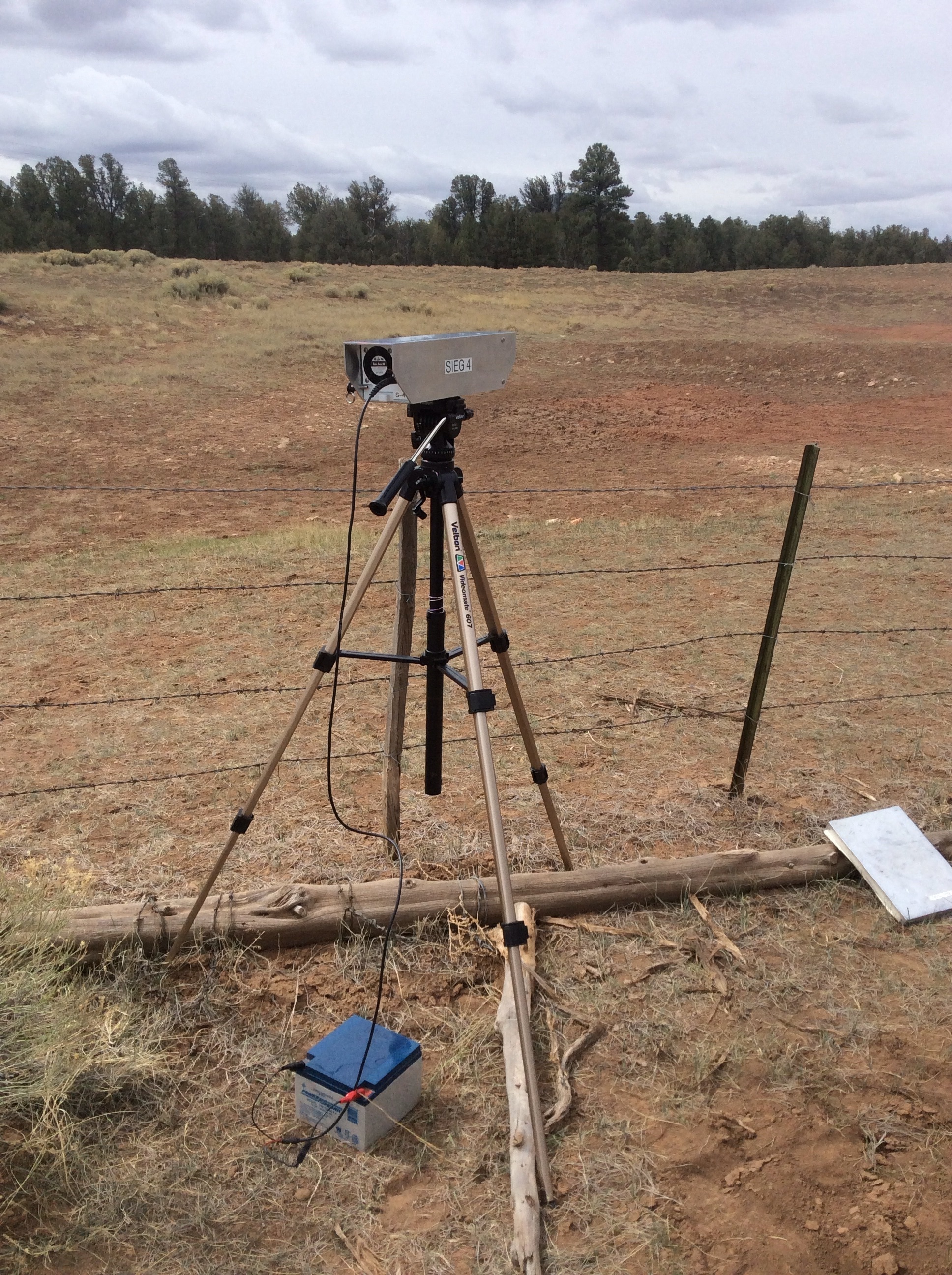  Active Dust sampler deployed on tripod attached to battery with barbed wire fence and bare ground in the near background, and pine trees on the horizon 