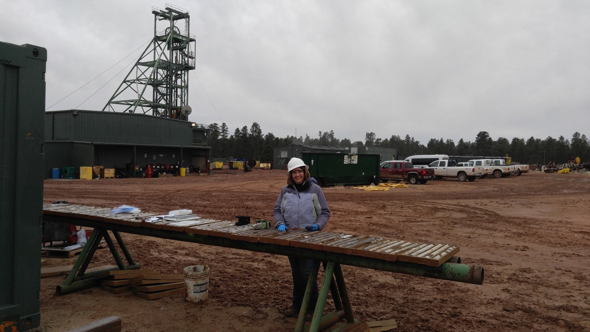  Scientist at long table covered with drill core. Mine buildings and head frame in the background. 
