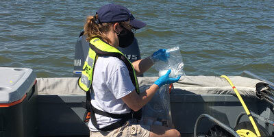 Female scientist in a boat, holding a plastic bag containing a plastic bottle recently filled with water from the lake for water quality analysis.  A water cooler for storing the samples is directly behind her.